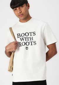 Fehér pamut póló, amelynek középpontjában merész fekete betűtípusban olvasható a "BOOTS WITH ROOTS" felirat, valamint egy kis fa grafika található, egy bézs válltáskával párosítva.