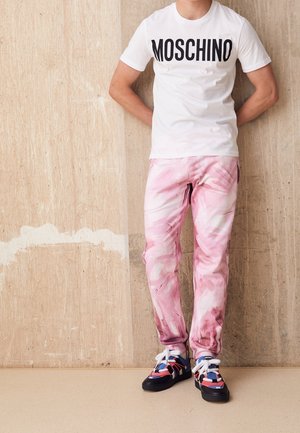 Biała koszulka z logo "MOSCHINO", spodnie w różowy marmurkowy wzór oraz wielokolorowe sneakersy z czerwonymi, niebieskimi i czarnymi akcentami.