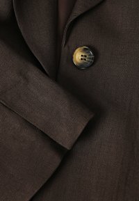 Tissu marron foncé à tissage fin montrant un gros plan d'une manche pliée et d'un grand bouton rond à deux trous.