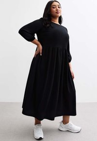 Robe midi noire à côtes avec un buste ajusté et une jupe évasée. Elle présente des manches trois-quarts et un col rond. Portée avec des baskets blanches.