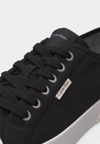 Μαύρο αθλητικό παπούτσι Calvin Klein με μαύρα κορδόνια, μεταλλικές θηλιές, λευκή σόλα και μικρή ετικέτα της μάρκας στο πλάι.