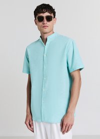 Hellblaues kurzärmeliges Hemd mit Stehkragen, strukturiertem Stoff und weißen Knöpfen. Trägt dunkle Sonnenbrille.