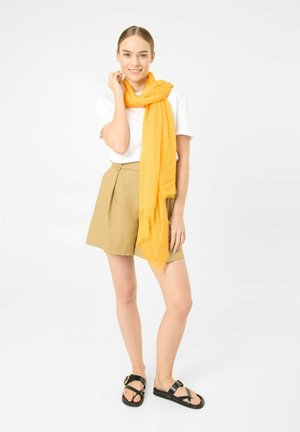 Jonge vrouw lacht, draagt een wit T-shirt, beige korte broek, gele sjaal en zwarte sandalen, staand tegen een effen witte achtergrond.