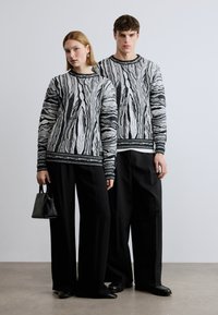 Pulls en maille texturée noir et blanc avec un motif tourbillonnant. Les modèles portent des pantalons noirs amples. L'un d'eux tient un petit sac à main noir.