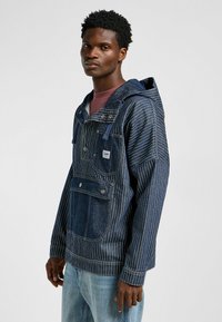 Die denimfarbene Kapuzenjacke hat vertikale weiße Streifen, eine Fronttasche und ein Markenpatch. Sie wird mit einem burgunderfarbenen Shirt und hellblauen Jeans getragen.