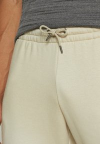 Beige sweatpants har et struktureret stof, elastisk talje med snor og metalspidser på snoren for holdbarhed.