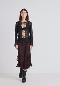 Top lung cu mânecă lungă, transparent negru, cu detalii legate, suprapus peste o fustă din franjuri plisați de culoare maronie, asortat cu bocanci negri până la glezne. Fundal neutru.