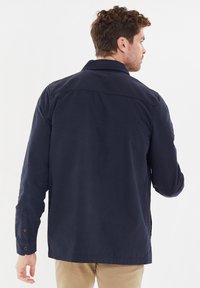 Marineblauwe jas met een gestructureerde kraag, knoopdetails aan de mouwen en een gladde stoftextuur. De achterkant toont stikseldetails en een ontspannen pasvorm.
