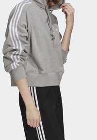 Grå kort hoodie med huva, med vita ränder längs ärmarna och en liten logotyp på bröstet. Tyget verkar mjukt och strukturerat.