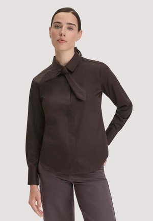 Junge Frau mit dunklem zurückgebundenem Haar, die eine dunkelbraune langärmelige Bluse mit Knotendetail am Kragen und passende Hose trägt.