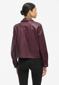 Veste en cuir bordeaux avec un design court, un col pointu et des manches longues avec des poignets à boutons. Texture lisse avec un léger brillant.