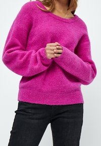 Magenta dzianinowy sweter o miękkiej fakturze, szerokim dekolcie i długich rękawach. Dół jest lekko prążkowany, noszony z czarnymi dopasowanymi dżinsami.