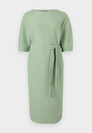 Femeie care poartă o rochie midi tricotată verde deschis cu mâneci trei sferturi, o curea în talie și sandale negre cu vârf deschis, stând în fața unui fundal alb.