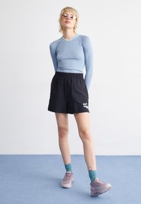 Jasnoniebieska bluzka z długim rękawem, czarne szorty z elastycznym pasem z logo, turkusowe skarpetki i jasnofioletowe buty sportowe na niebieskim tle.