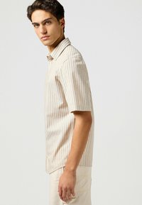Camicia a maniche corte con bottoni in tessuto beige chiaro, con strisce verticali color crema, dotata di colletto a punta e fessure laterali per la circolazione dell'aria.