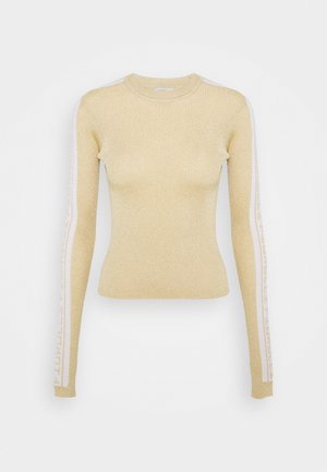 Pull ajusté beige à manches longues avec un léger scintillement et des rayures blanches portant un texte courant le long des deux manches.