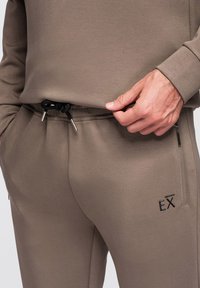 Gros plan sur une personne ajustant le cordon de serrage d'un pantalon de survêtement taupe avec une poche zippée et un logo noir "EX" sur la cuisse.