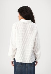 Blusa blanca de manga larga con un patrón texturizado de diamantes, con cuello tipo camisa y dobladillo ligeramente redondeado.