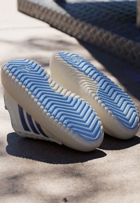 Bijele sportske tenisice s teksturiranim plavim đonom i tri ljubičaste pruge sa strane, koje imaju uzorak valova i elegantan dizajn.