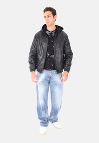Giovane uomo che indossa una giacca in pelle nera con cappuccio, un maglione nero con motivo, jeans chiari sbiaditi e sneakers bianche, in piedi contro uno sfondo semplice.