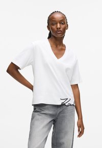 T-shirt bianco in cotone con scollatura a V e maniche corte, con un dettaglio della firma ricamato in nero sull'orlo. Abbinato a jeans grigi.