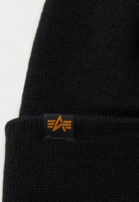 Gorro de lana negro con un acabado texturizado y una etiqueta de tela negra que presenta un logotipo naranja y líneas decorativas. Forma compacta para mayor calidez.