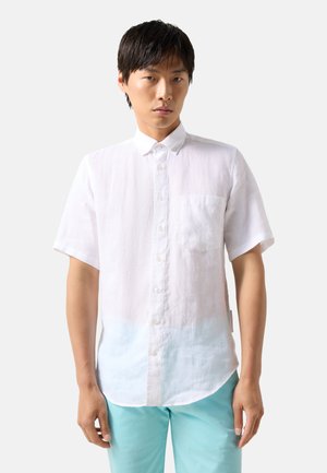 Mann trägt ein weißes, kurzärmeliges Hemd mit Brusttasche und hellblaue Hose, steht vor einfachem Hintergrund.