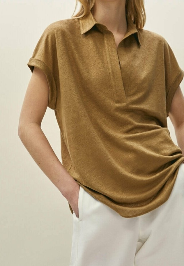 Femme portant une chemise marron ample à manches courtes avec col et un pantalon blanc, une main dans la poche, fond neutre.