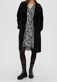 Long manteau noir texturé porté sur une robe à rayures zèbre noir et blanc. Collants noirs et bottines noires épaisses. Col montant et fermeture à boutons.