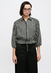 Πουκάμισο με κουμπιά σε ασπρόμαυρο καρό, μανίκια τριών τετάρτων και εφαρμοστή μέση. Συνδυάζεται με μαύρο παντελόνι ραμμένο.