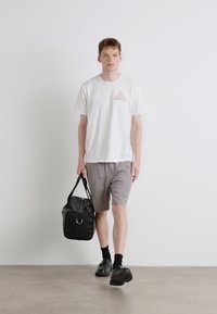 White t-shirt with a patterned triangle on the chest, gray shorts, black shoes, and carrying a black duffel bag with a circular detail.