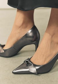 Spitz zulaufende High Heels aus metallisch grauem Leder mit einem Knoten-Detail auf dem Blatt und einem Stiletto-Absatz, der eine glatte, glänzende Oberfläche zeigt.