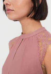 Ροζ μπλούζα με ψηλό λαιμό, που διαθέτει δαντελένιες λεπτομέρειες στους ώμους και άνοιγμα-κλειδί στο μπροστινό μέρος. Μαλακό, υφασμάτινο ύφασμα με υφή.