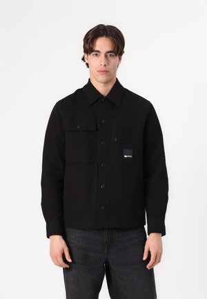 Black button-up jacket made of cotton with two front pockets. Features a pointed collar and a logo patch on the left chest.