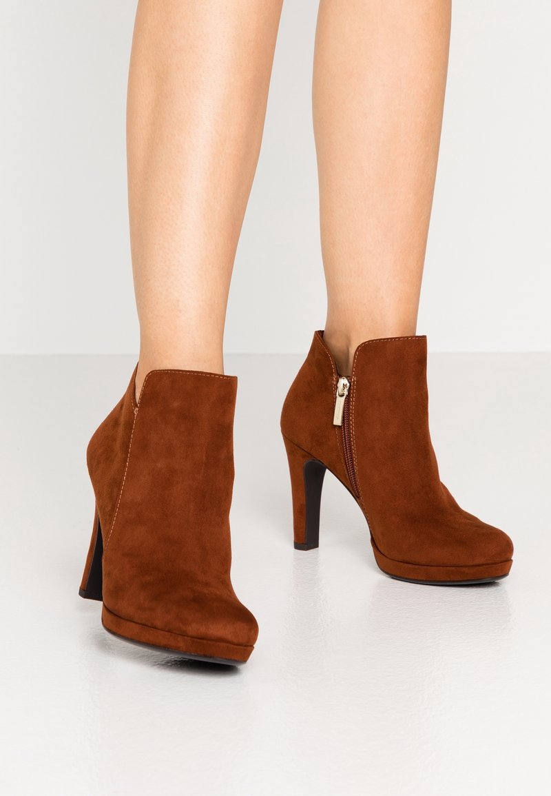 Bottines en suède marron avec fermeture éclair latérale et talons hauts portées sur des jambes nues sur un fond uni.