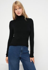 Pullover preto de gola alta feito de um tecido leve e macio. Apresenta mangas longas e um design ajustado, combinado com jeans azuis de cintura alta.