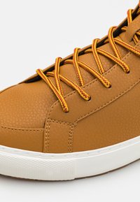 Zapatilla de piel sintética marrón con superficie texturizada, cordones amarillos y rojos en contraste y suela de goma blanca. Presenta detalles con costuras.