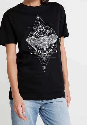 Sort t-shirt med et detaljeret hvidt mønster af en natsværmer centreret i geometriske former og symboler, båret af en person i lyseblå jeans.