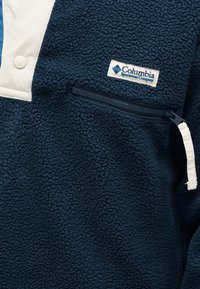 Μπουφάν από φλις σε ναυτικό μπλε με λευκά κουμπιά πατιλέτα, οριζόντια τσέπη με φερμουάρ και ετικέτα της εταιρείας Columbia Sportswear Company στο στήθος.