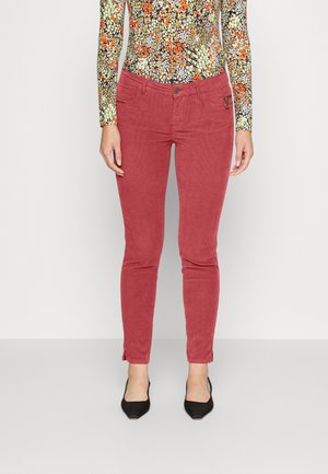 Czerwone spodnie o wąskim kroju, lekko wyblakłe, noszone z długim rękawem w kwiaty i czarne buty na spiczasty nosek na jednolitym jasnym tle.