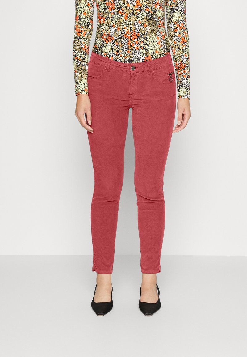 Pantalon rouge délavé coupe slim porté avec un haut à fleurs à manches longues et des chaussures noires à bout pointu sur un fond clair uni.