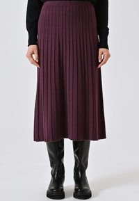 Donna con una gonna midi viola a costine, maglietta nera a maniche lunghe e stivali in pelle neri fino al ginocchio, in piedi davanti a uno sfondo uniforme.