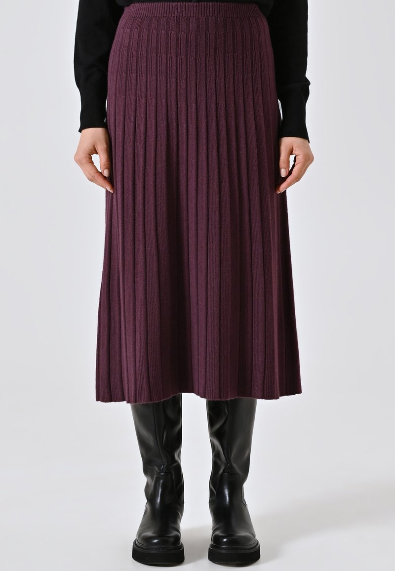 Donna con una gonna midi viola a costine, maglietta nera a maniche lunghe e stivali in pelle neri fino al ginocchio, in piedi davanti a uno sfondo uniforme.