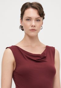 Top senza maniche color bordeaux con scollo a cowl. Realizzato in tessuto morbido, presenta una texture liscia e un design aderente.