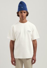 Camiseta blanca de algodón con mangas cortas, que presenta un pequeño logotipo impreso en el pecho. Combinada con un gorro de punto azul.