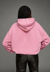 Pull à capuche rose avec une finition texturée, doté d'une grande capuche, d'accents cloutés le long des épaules, accompagné de pantalons en cuir noirs.
