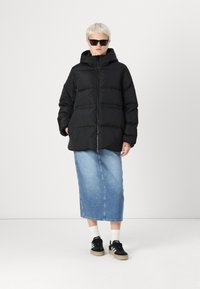 Juodas pūkinis švarkas su gobtuvu ir užtrauktuku, su išsiuvinėta dizaino struktūra. Dėvimas virš ilgo džinsinio sijono ir derinamas su juodais sportbačiais.