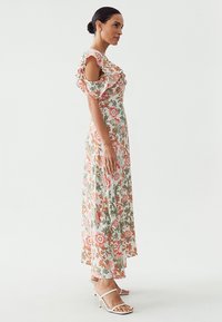 Femme dans une robe maxi à fleurs, décolletée et avec des manches volantes, se tenant de côté et portant des sandales à talons blancs, sur fond uni.