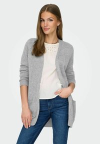Cardigan gris clair à manches longues, à ouverture devant et avec des poignets côtelés. Porté sur une blouse blanche avec des détails en broderie, assorti à un jean bleu.