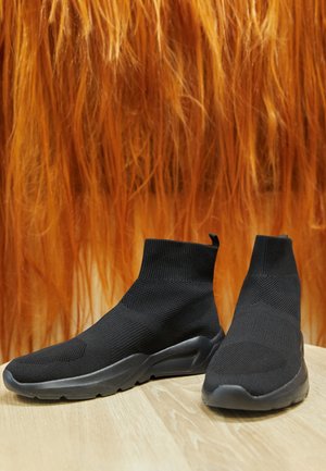 Paire de baskets chaussettes noires en tricot avec semelles épaisses sur un sol en bois clair et un fond texturé orange.